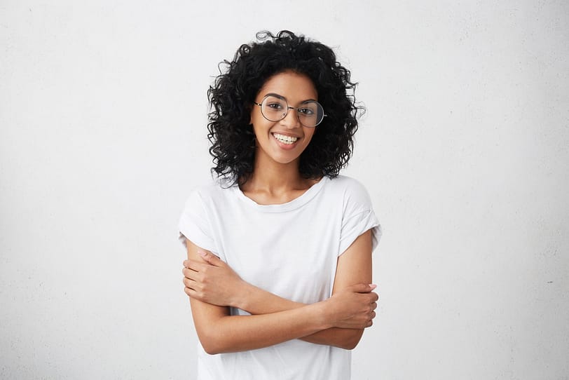 Indoor Shot Of Beautiful Happy African American Student Girl Standing Isolated In White Studio Smiling Cheerfully, Keeping Her Arms Folded, Relaxing Indoors After Morning Lectures At University
